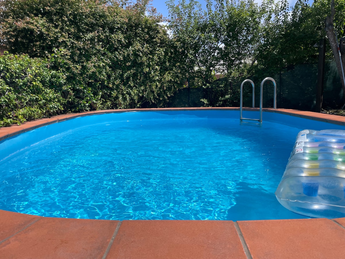 A private pool is set in a well-kept garden area, featuring clear blue water and a smooth tile edge. A pool ladder provides easy access, while a floating lounge adds a touch of relaxation. Lush green foliage surrounds the pool, offering privacy.