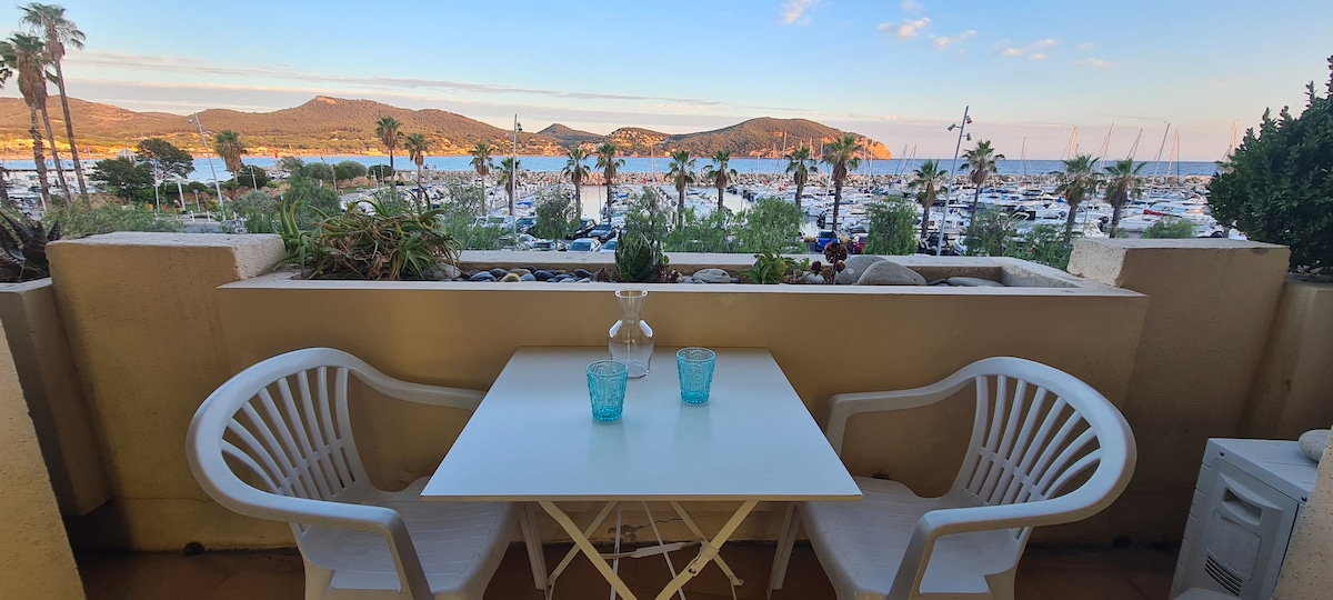 A covered terrace features a white table surrounded by two chairs, overlooking a scenic view of the sea, palm trees, and boats in the harbor. Natural light illuminates the space, enhancing the peaceful surroundings.