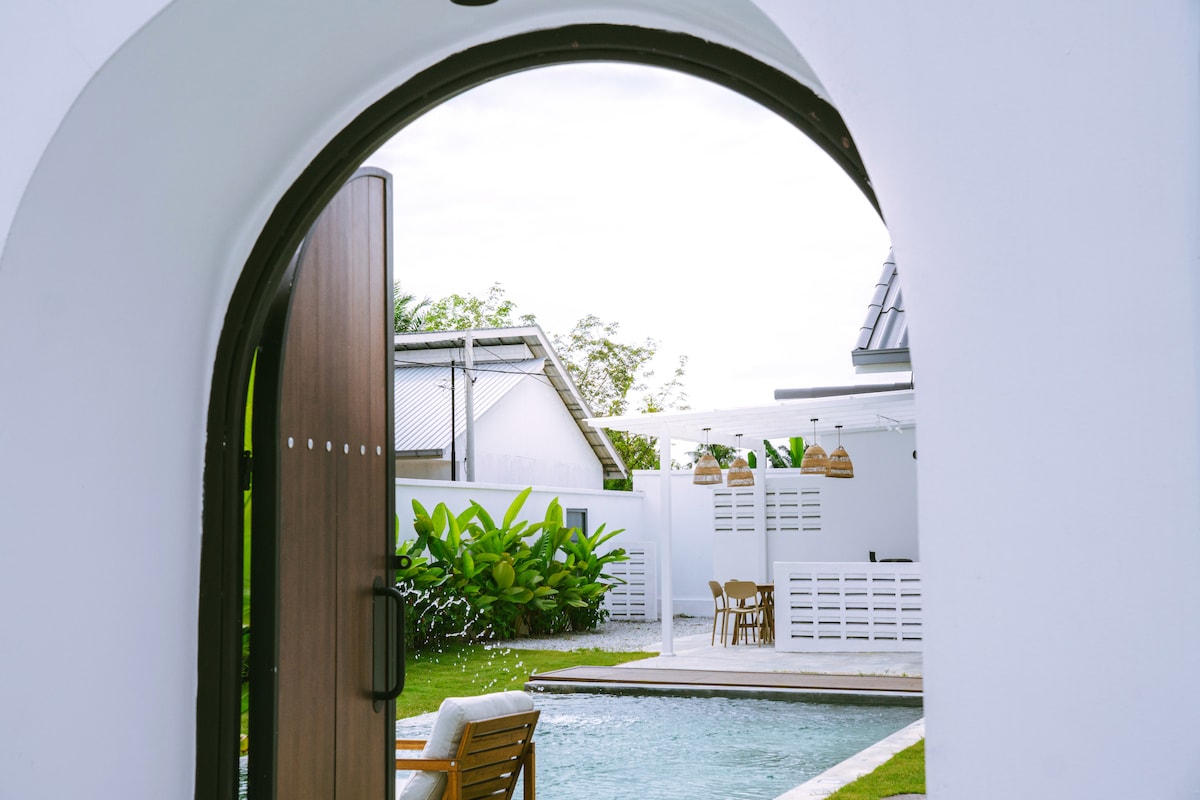 A graceful arched doorway frames a view of the private pool and outdoor dining area. Lush greenery surrounds the space, complementing the clean lines of the modern villa. Subtle water movement in the pool reflects the tranquil atmosphere of the retreat.