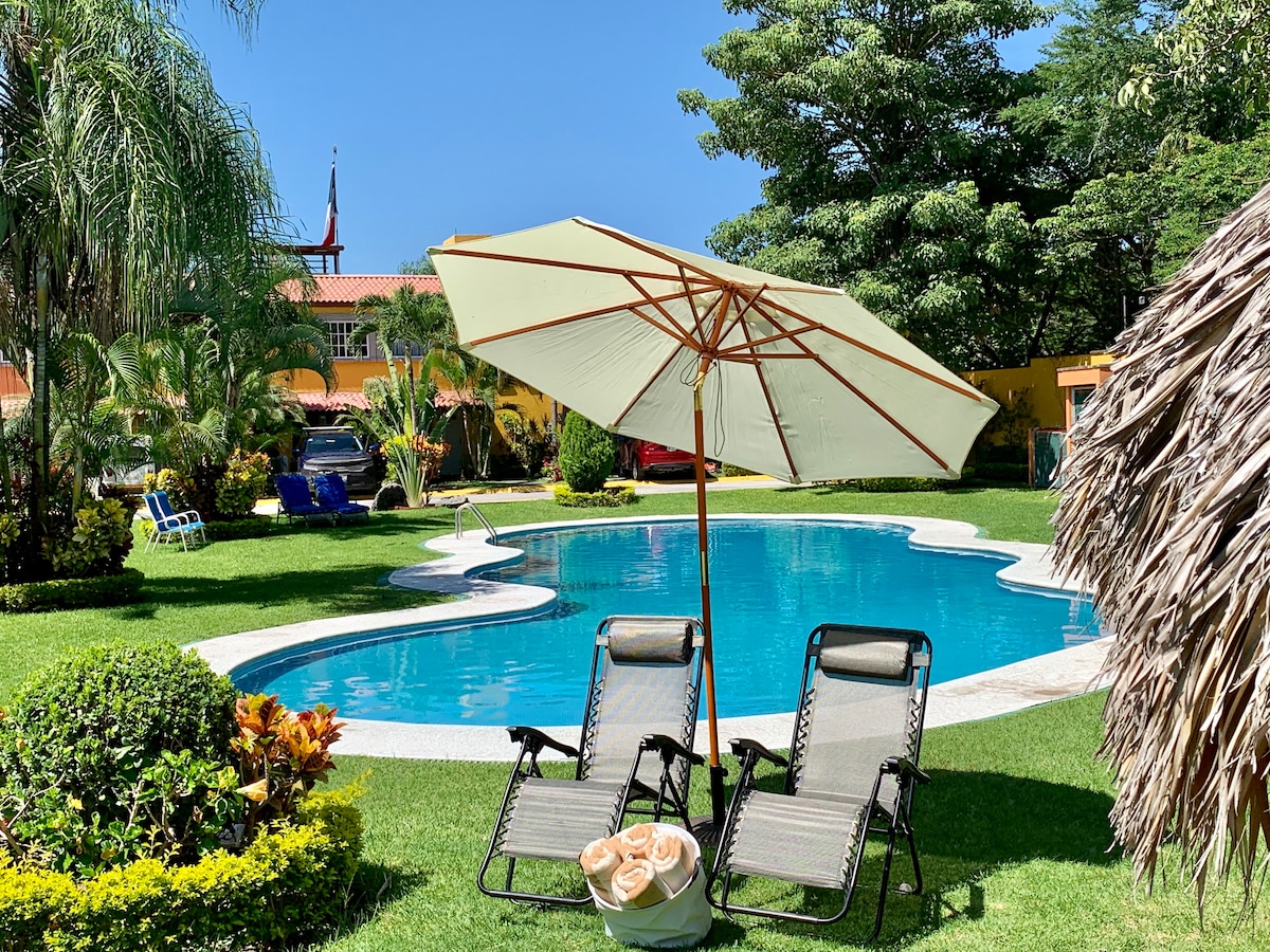 A refreshing pool area is framed by lush greenery and tropical plants. Two lounge chairs are positioned beneath a large umbrella, providing shade. A round pool features a curvy shape, inviting relaxation in a sunny environment. Nearby, vibrant flowers add natural beauty.
