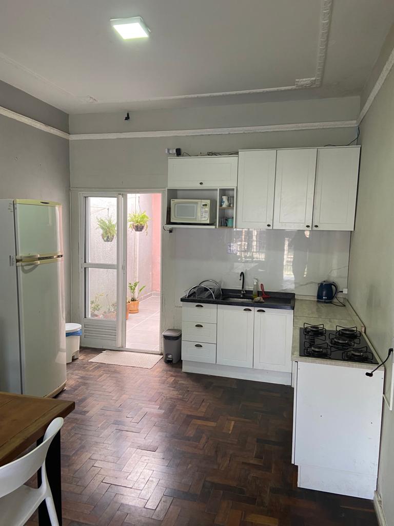 A modern kitchen features white cabinetry and a tiled backsplash, equipped with a refrigerator, microwave, and electric kettle. A wooden dining table offers space for meals, while a dual-burner stove is located on the countertop. Natural light enters through glass doors leading to an outdoor area.