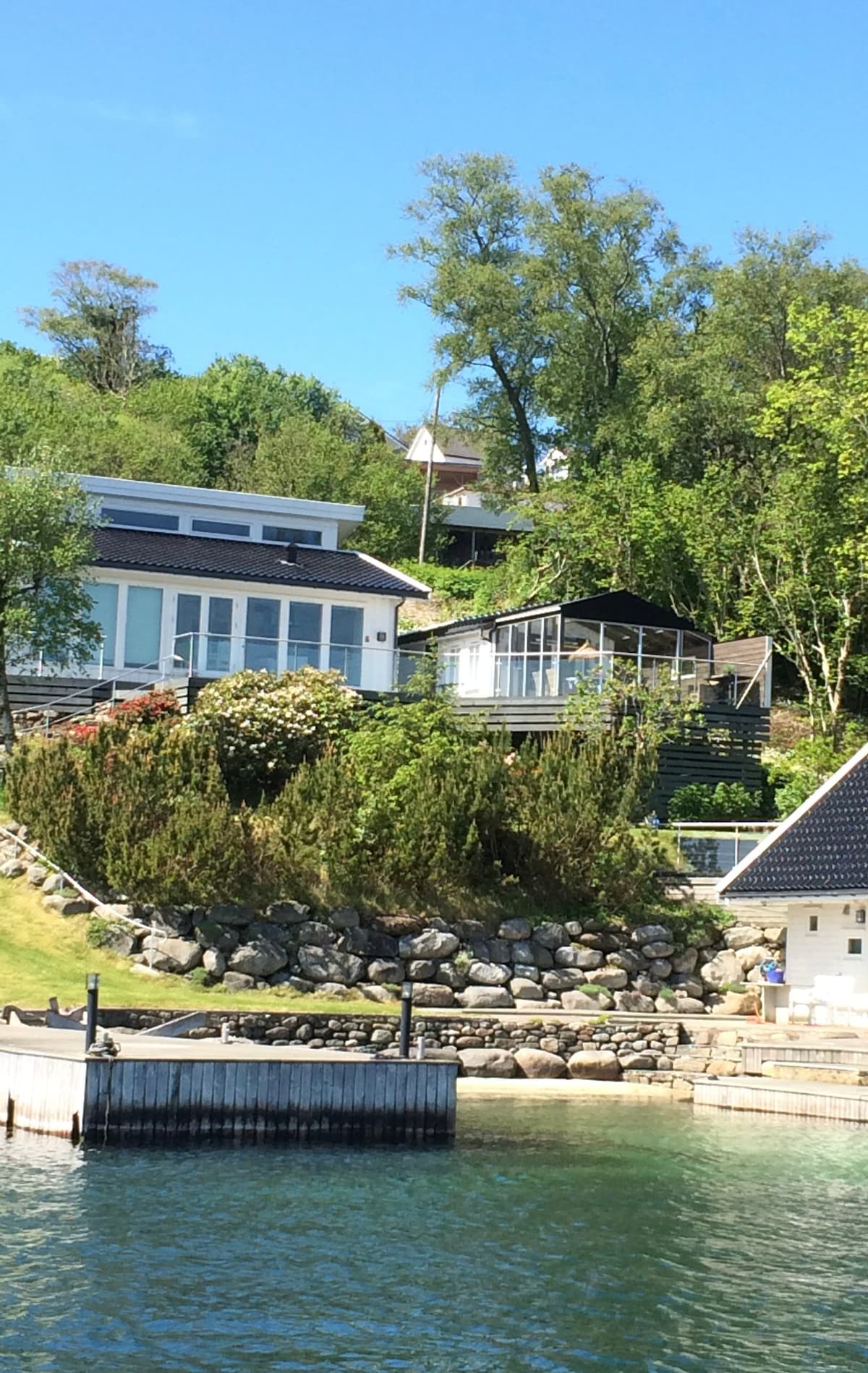 A charming modern cabin is set in a natural landscape, surrounded by trees and lush foliage. A cozy patio with glass walls is visible, offering views of the serene water. The cabin is situated just above a private dock, enhancing the scenic waterfront appeal.
