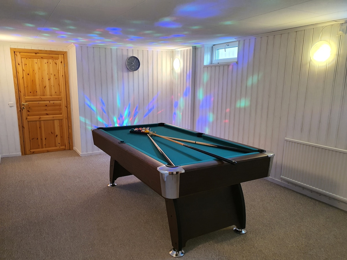 A billiard table is centrally located in a well-lit game room, featuring colorful lights reflecting off the walls. A wooden door and clock are visible in the background, contributing to the welcoming environment.