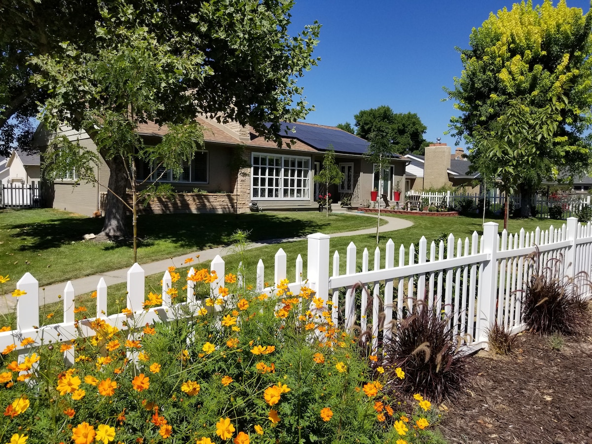 The exterior of a charming house is framed by a white picket fence and colorful flower beds featuring bright yellow blooms. A well-kept lawn stretches in front, complemented by mature trees. The sunny sky creates a warm ambiance in this residential neighborhood.