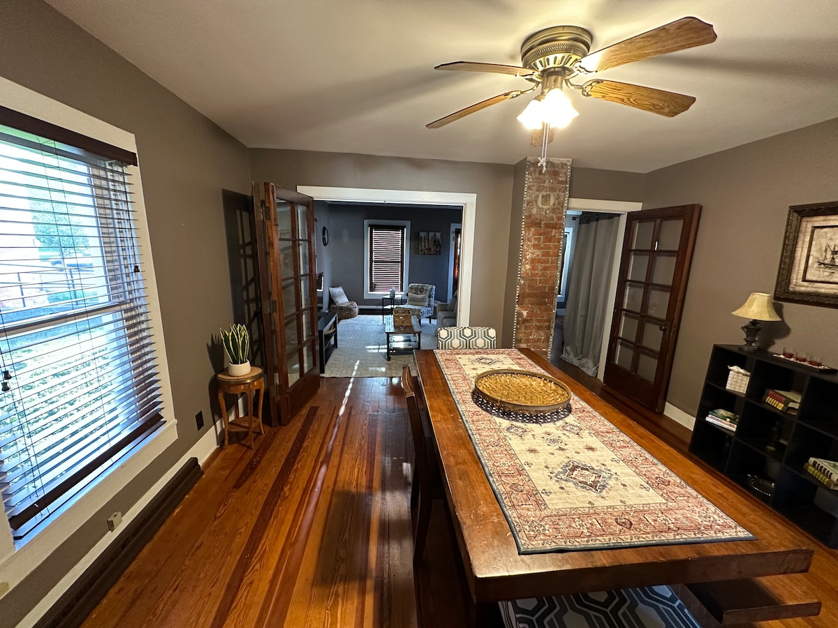 The dining room showcases a large wooden table with a decorative centerpiece, complemented by a ceiling fan. Natural light filters through the windows, illuminating the wood flooring and the inviting atmosphere. French doors create an open flow to the adjoining living area.