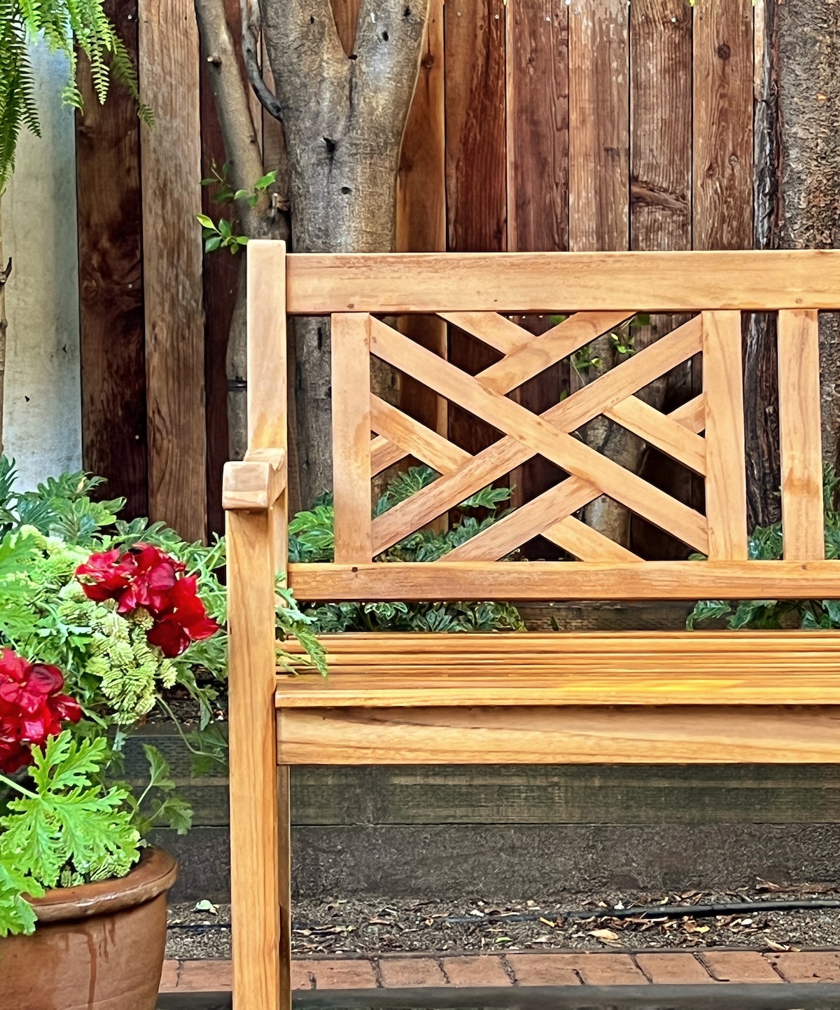 A wooden bench features a geometric backrest design, positioned nearby vibrant red flowers in a terracotta pot. Surrounding foliage creates a natural, inviting atmosphere in the garden.