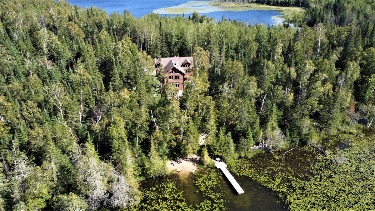 An aerial view captures the lodge nestled among lush trees, with Jack The Horse Lake in the background. A private dock extends into the lake, surrounded by vibrant greenery and lily pads, creating a serene natural setting.