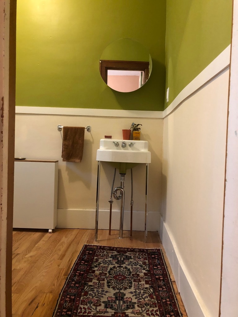 A small bathroom space features a vintage sink with elegant legs and a round mirror above it. A towel hangs on a nearby rack. The walls are painted in a bright green, and a patterned rug covers part of the wooden floor.