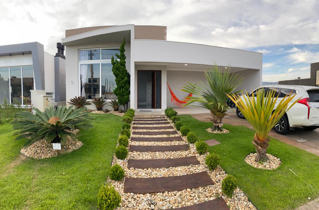 The exterior of the house features a well-kept garden with a variety of palm plants and decorative stones lining the pathway. A contemporary facade is showcased, with large glass windows and a welcoming entrance, complemented by a colorful hammock hung nearby.