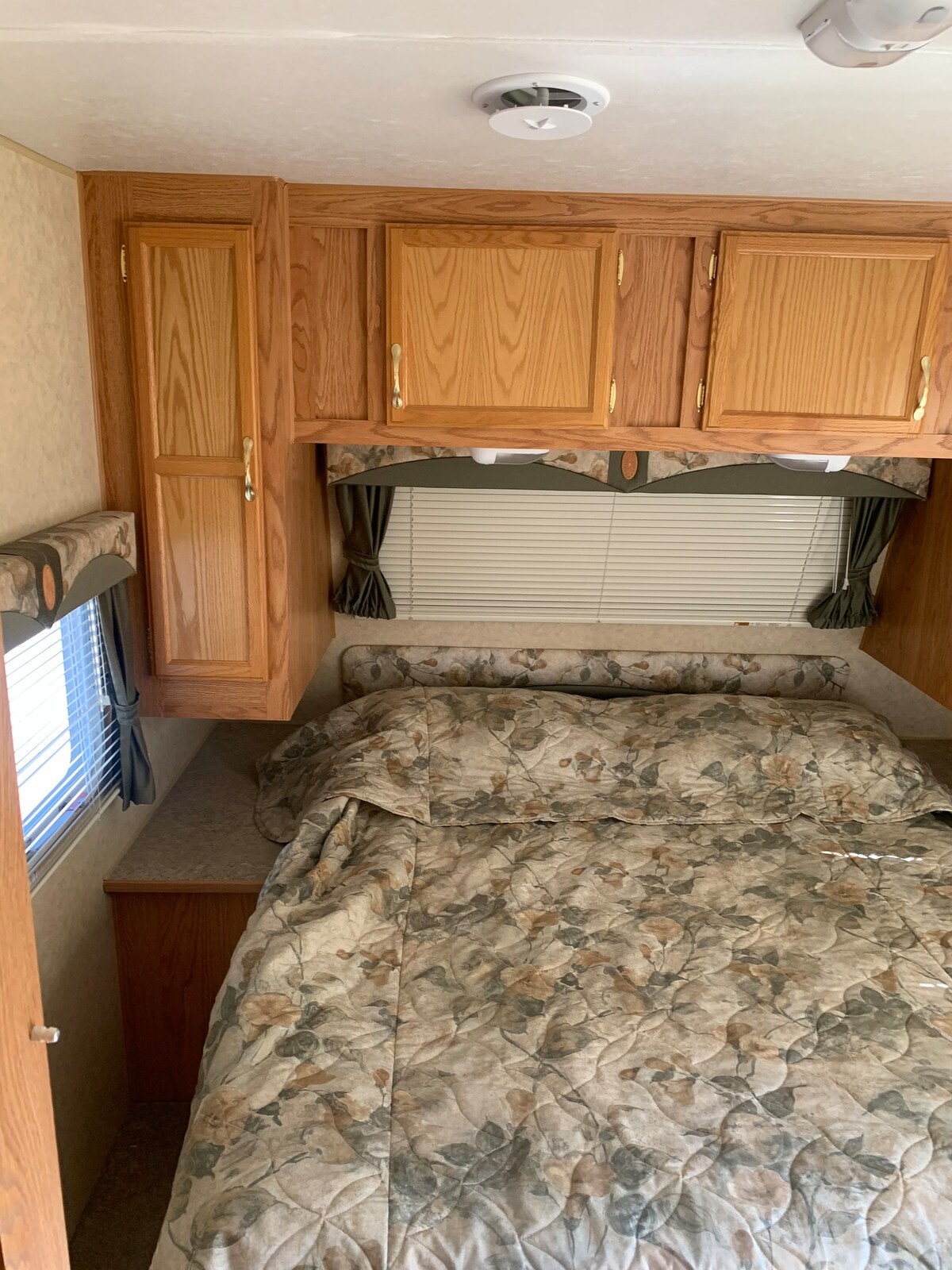 A cozy sleeping area features a spacious bed adorned with a floral-patterned comforter. Wooden cabinetry above provides additional storage, while a softly draped window treatment enhances the light filtering in from the nearby window.