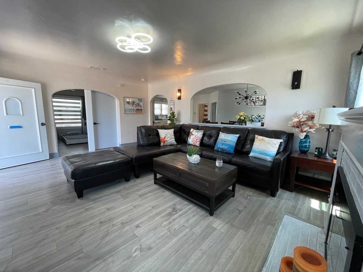 A spacious living room features a black sectional sofa with coordinating cushions, and a wooden coffee table at its center. Light-colored walls complement the wooden flooring. Doorways lead to other areas of the cottage, and sunlight brightens the space through large windows.