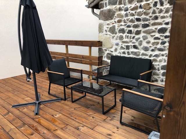 An outdoor wooden terrace features a seating arrangement consisting of two chairs, a sofa, and a glass-topped coffee table. A large umbrella stands nearby, and the rustic stone wall adds texture to the space. The wooden flooring imparts a warm and natural feel.