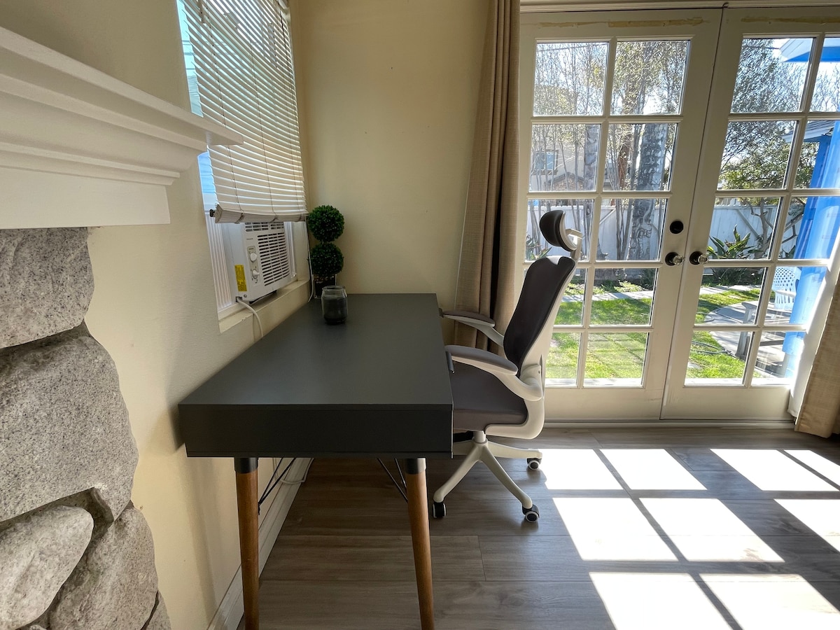 A workspace is shown, featuring a modern desk with wooden legs and a sleek surface. A comfortable office chair is positioned at the desk. Light enters through the French doors, illuminating the room and providing views of the private courtyard outside.