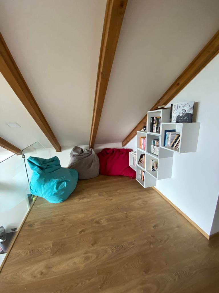 A bright corner of the upstairs chill-out area features colorful bean bags in shades of pink and blue, arranged on a warm wooden floor. A modern wall shelf holds neatly organized books, complementing the slanted ceiling with exposed wooden beams.