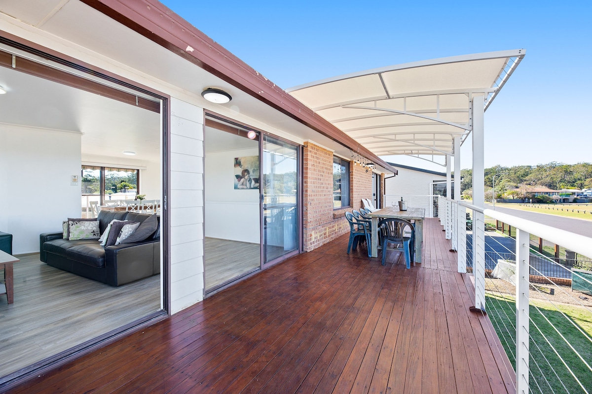 A spacious balcony with a wooden deck is partially covered, featuring a dining table and chairs. Glass doors lead into the living area. Scenic views of greenery are visible in the background, providing a tranquil outdoor space for relaxation.