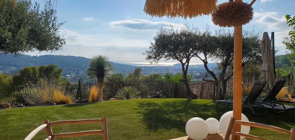 A garden area features a lush lawn with a distant view of the bay. Sun loungers are positioned under olive trees, and a thatched umbrella provides shade. Decorative white orbs add a modern touch to the outdoor space, enhancing the serene ambiance.