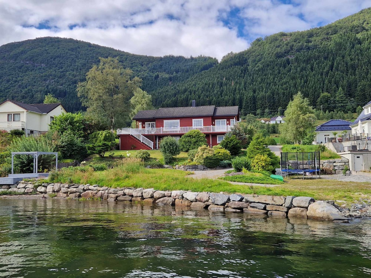 A spacious property is situated by the water, featuring a well-maintained garden with varied greenery and stone landscaping. The two-story red house is framed by mountains and trees. A trampoline and dock can be seen in the yard, enhancing the outdoor activity options.