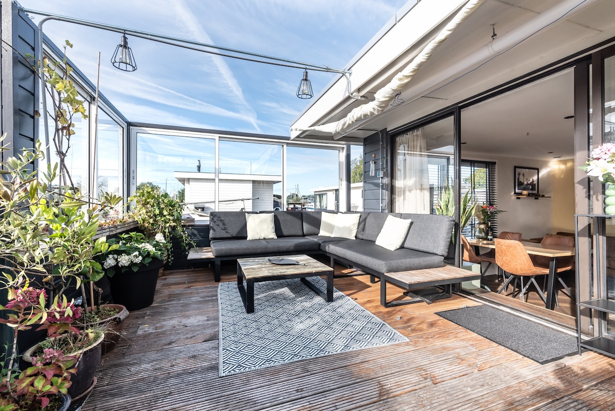 An outdoor terrace features a spacious seating area with gray cushions and a wooden coffee table. Surrounded by potted plants, the space is bathed in natural light, with large glass panels providing an unobstructed view. A cozy atmosphere is suggested with decorative elements and soft textures.