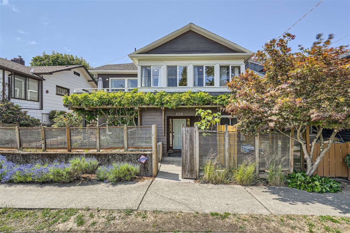 The front view of the home displays a welcoming entrance framed by greenery and colorful plant life. The exterior features a mix of wood and modern detailing, with large windows allowing natural light to fill the interior. A pathway bordered by vibrant flowers leads up to the entrance.