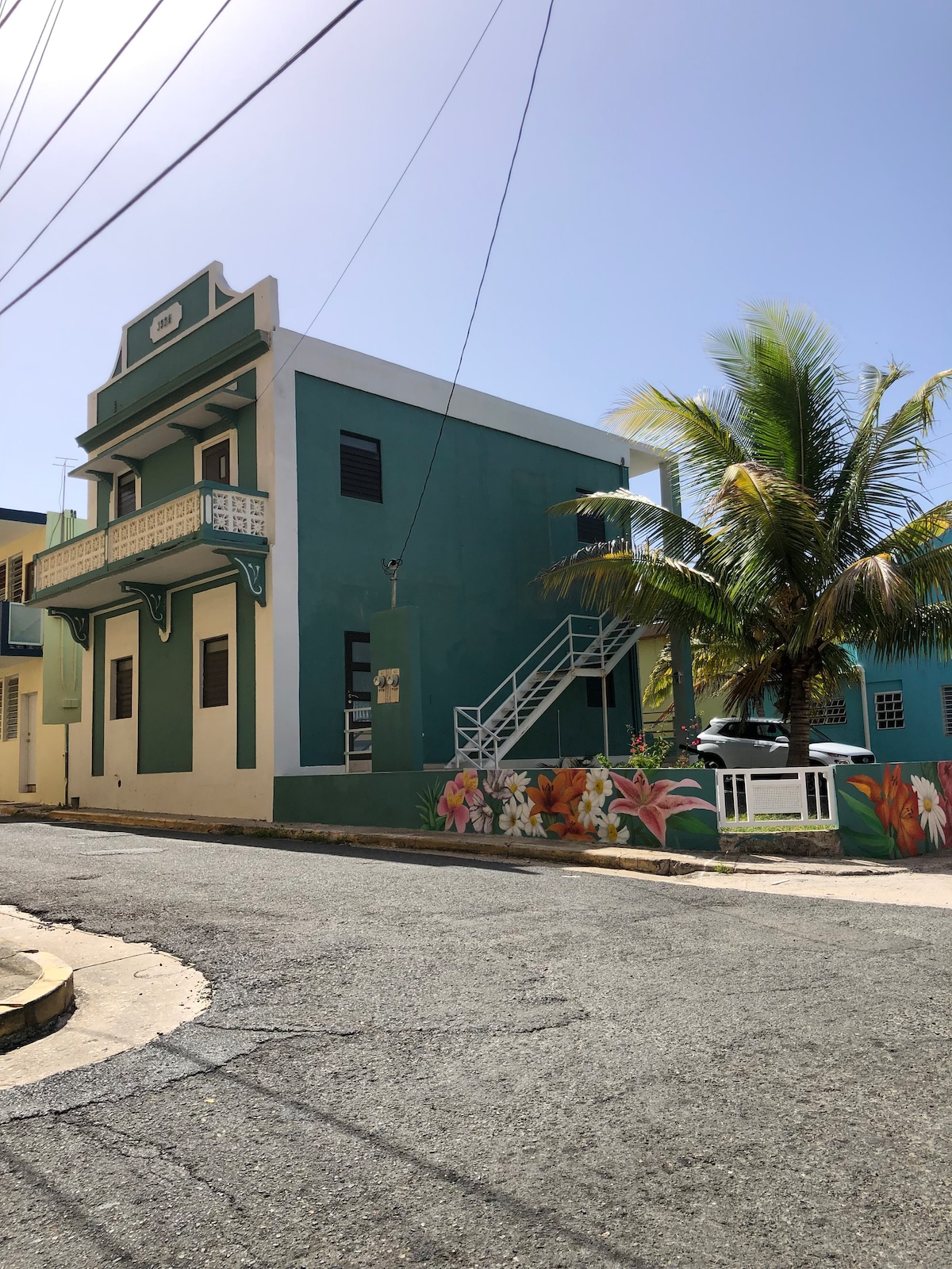 A vibrant, two-story building showcases a teal exterior, adorned with decorative white trim. A staircase leads to the second floor, while tropical palm trees and a colorful mural of flowers embellish the front. Power lines are visible overhead against a clear blue sky.