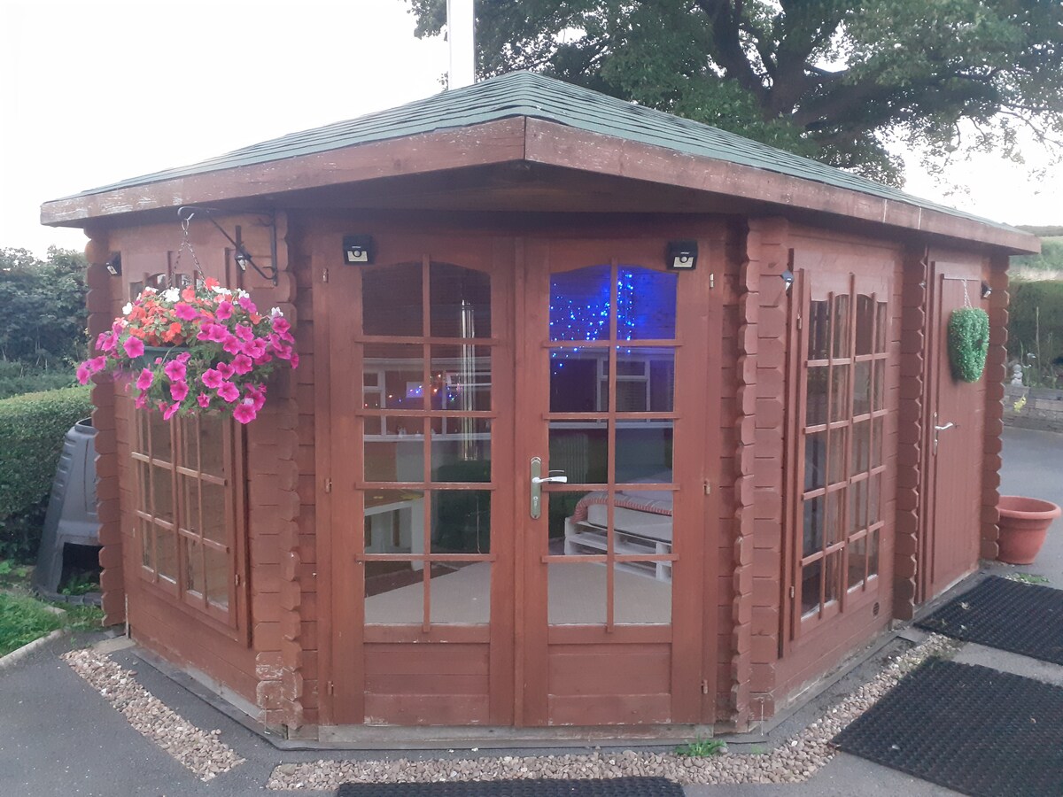 A wooden cabin featuring large glass doors and multiple windows, allowing natural light to fill the interior. Bright flower baskets hang at the entrance, and a cozy porch area is visible, providing a welcoming outdoor space surrounded by greenery.