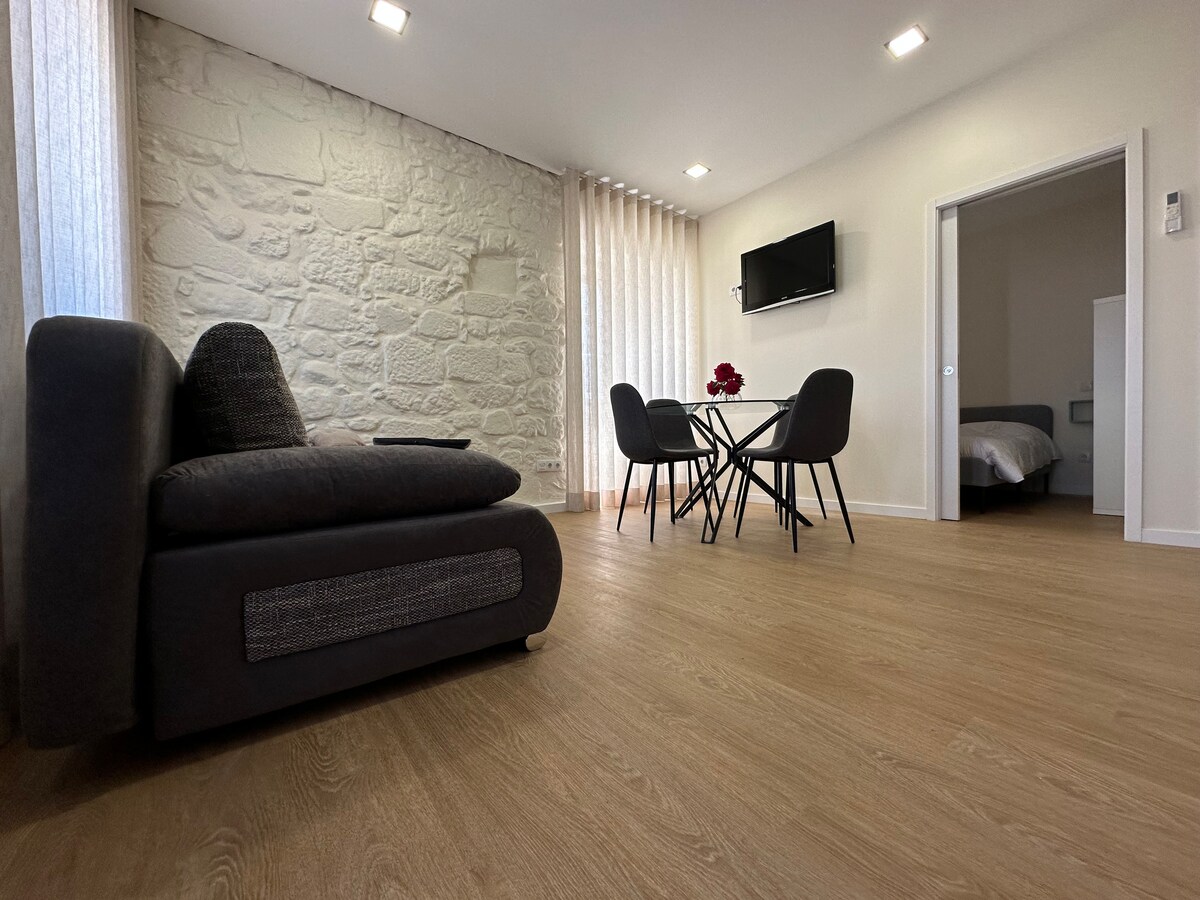 A modern living area is featured with a comfortable sofa and a small dining table surrounded by four chairs. A television is mounted on the wall, and natural light filters through sheer curtains beside a textured stone wall. A doorway leads to a bedroom in the background.