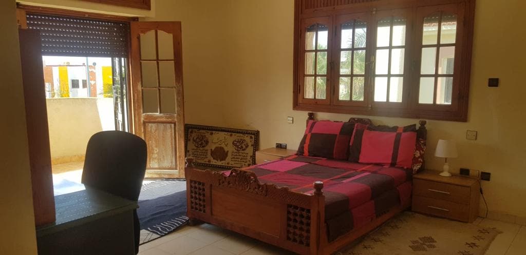 A well-furnished bedroom features a spacious bed adorned with a colorful blanket. Light streams in through large windows, offering views of the outdoors. A wooden desk and chair enhance the workspace, while decorative accents add character to the room.