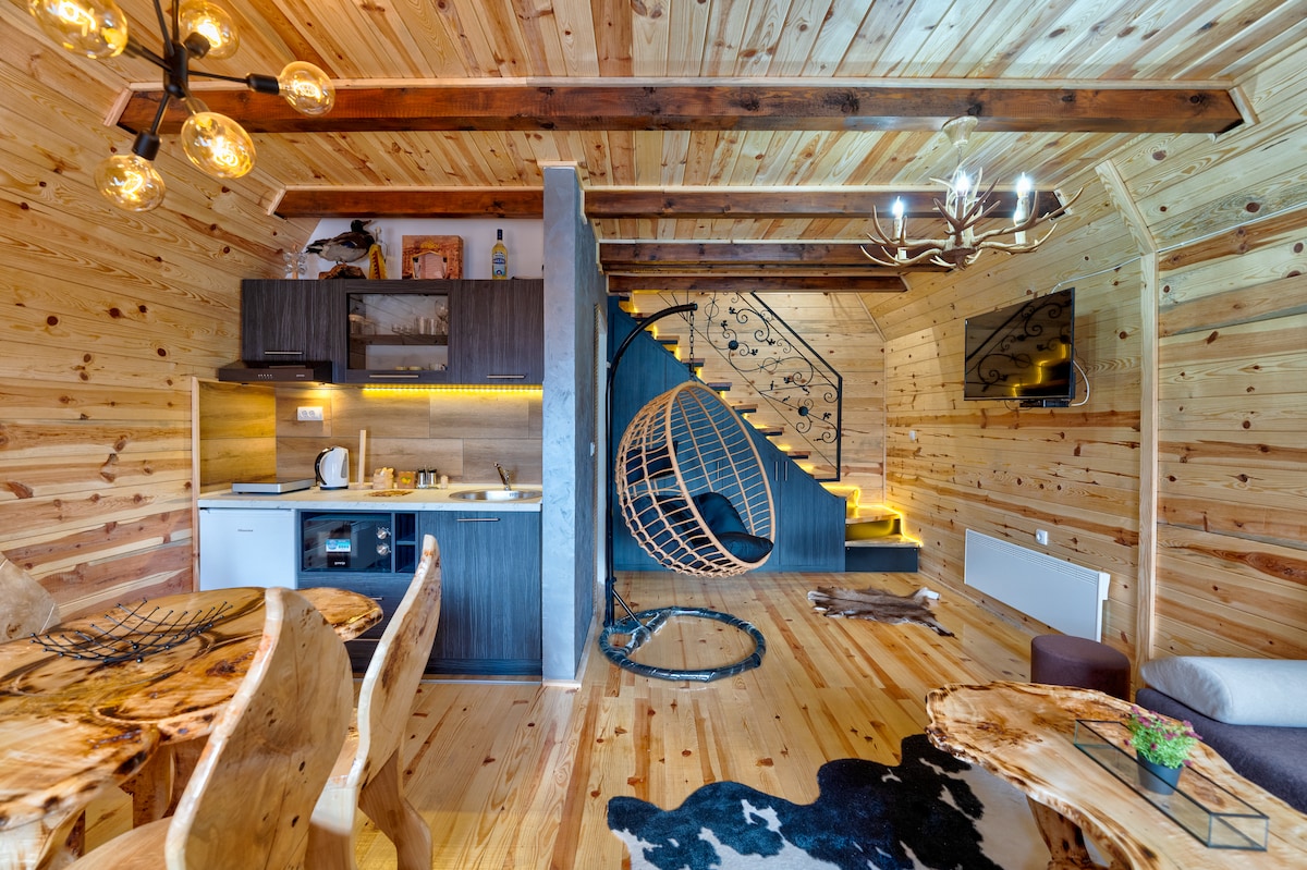 A warm wooden interior features an open layout with a kitchen area, dining table, and a staircase leading to the upper level. Unique hanging seating is present, alongside a textured rug on the floor. Soft lighting enhances the inviting atmosphere.