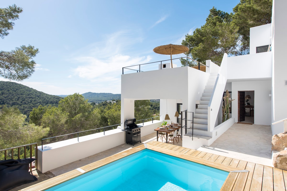 An outdoor area showcases a private swimming pool surrounded by wooden decking. A staircase leads to a spacious terrace adorned with lounge furniture and an umbrella, offering views of the greenery and hills. A BBQ grill stands nearby, enhancing the outdoor dining experience.