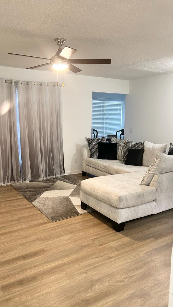 A comfortable sitting area features a light-colored sectional sofa adorned with dark accent pillows. A patterned area rug lies beneath, complemented by large, sheer curtains that allow natural light to filter through. A ceiling fan adds a touch of elegance to the space.