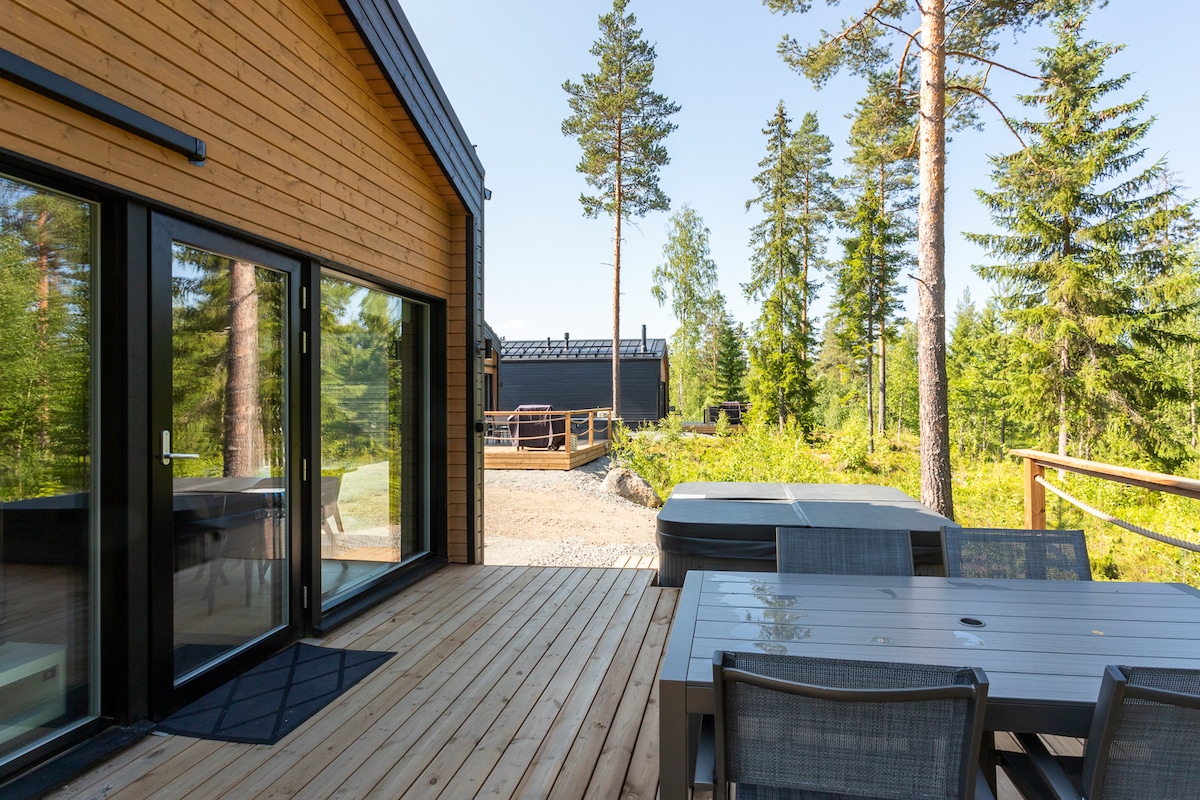 A wooden terrace is shown, furnished with a rectangular table and several chairs. A hot tub is visible beside the table, surrounded by trees. The exterior walls exhibit a natural wood finish, and large glass doors reflect the outdoor scenery.