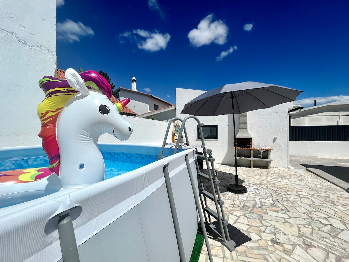 An above-ground pool is presented with a colorful unicorn float. A gray umbrella is set beside the pool, providing shade. A barbecue area is visible in the background, while the surrounding patio features natural stone flooring and a clear blue sky above.