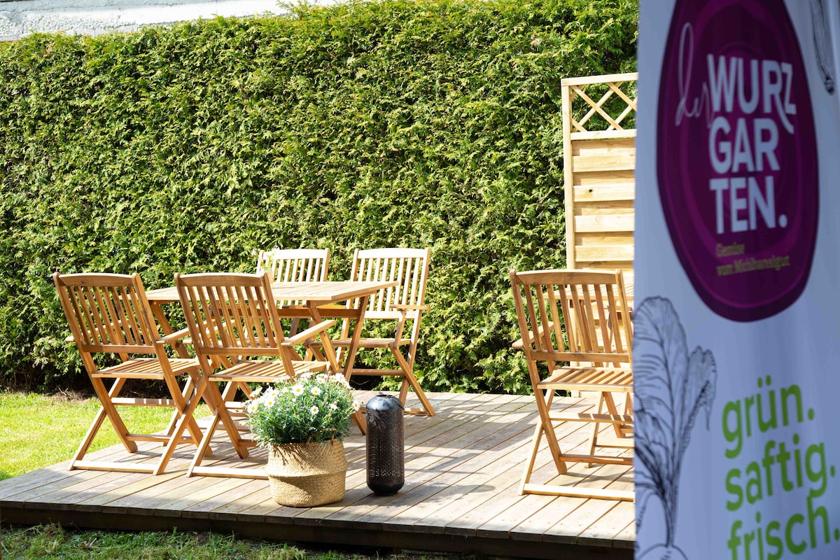 An outdoor seating area is visible, featuring a wooden deck with a rectangular table and multiple folding chairs arranged around it. A potted plant with white flowers adds a natural touch. A tall hedge provides privacy, creating a serene environment for relaxation.