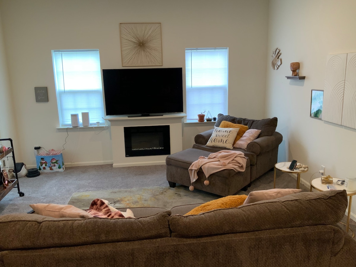 A cozy living area features two plush sofas positioned around a central coffee table. Light from two windows enhances the space, while a modern television is mounted above a sleek electric fireplace. Soft decorative elements, such as cushions and throw blankets, add a welcoming touch.