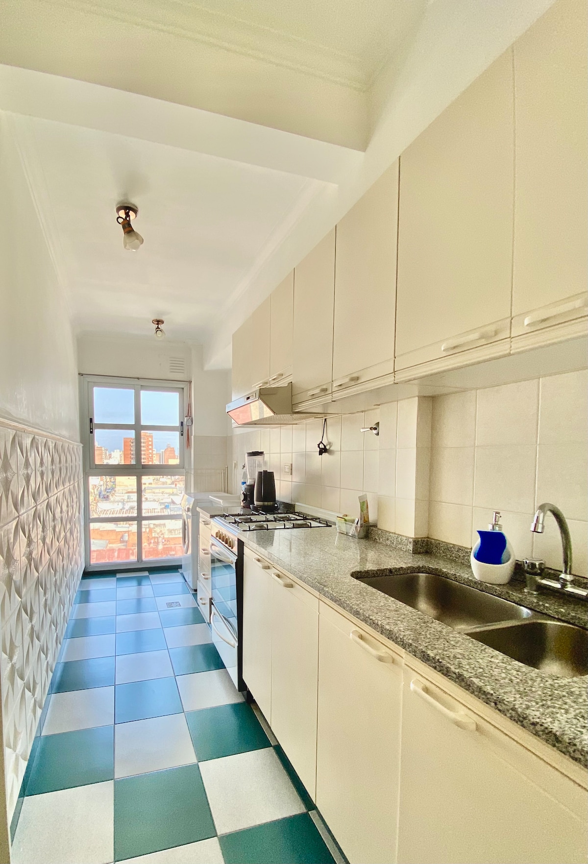 A modern kitchen features light-colored cabinetry and a granite countertop. Stainless steel appliances include a gas stove and refrigerator. The floor showcases a pattern of green and white tiles, while a large window provides natural light and a view of the cityscape beyond.