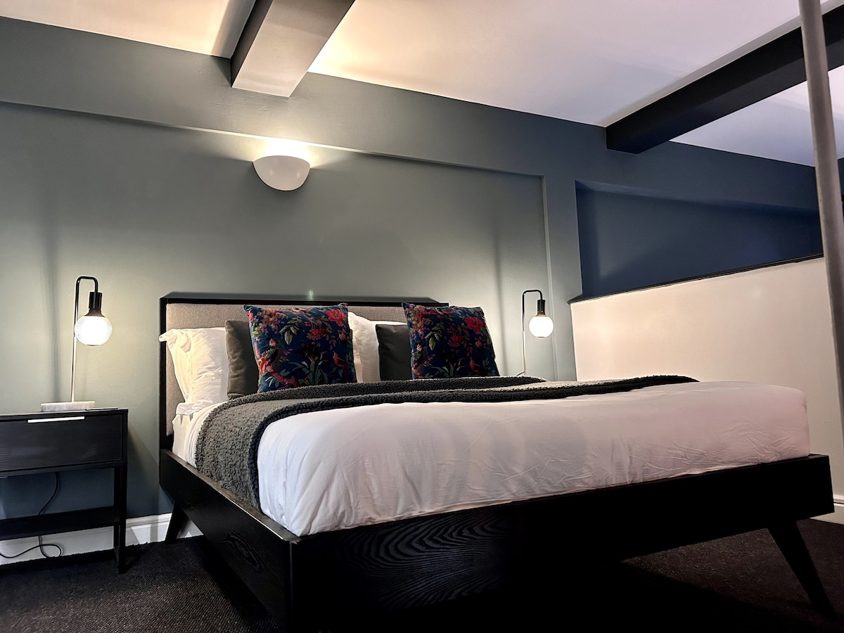 A spacious sleeping area features a large bed with a dark wooden frame and soft bedding. Two decorative pillows add color to the bed. Two bedside lamps provide soft lighting against a dark green accent wall.