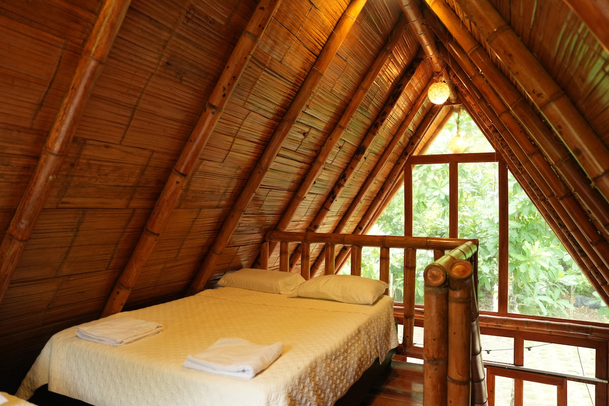 A cozy sleeping area is featured under a sloped bamboo ceiling. A spacious bed is dressed in a light-colored coverlet, flanked by freshly laundered towels. Large windows reveal greenery outside, enhancing the serene ambiance of the room.