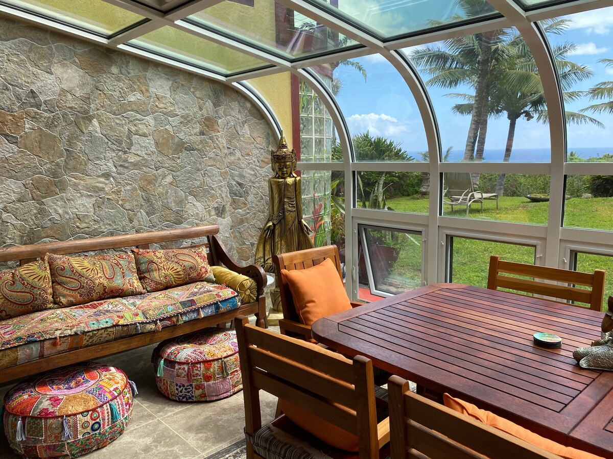 A sunlit solarium features a stone wall and large windows with views of the lush garden and ocean beyond. Colorful cushions and patterned poufs adorn a wooden couch, while a dining table invites gatherings. Tropical palm trees are visible through the glass enclosure.