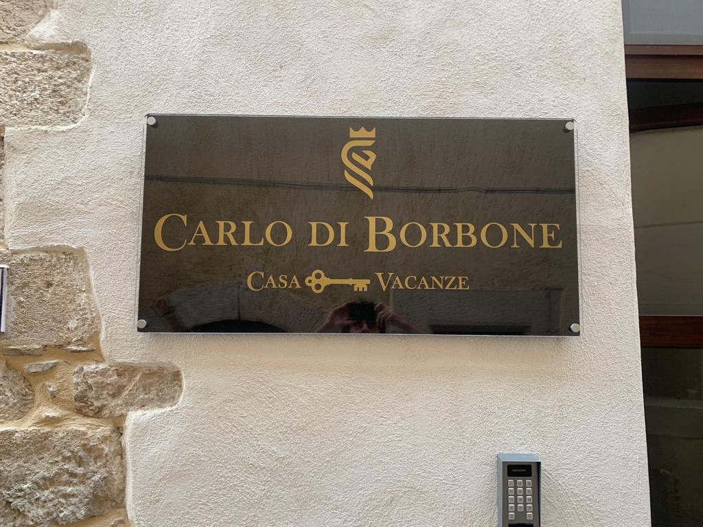 A polished black sign displays the name 'Carlo di Borbone' in golden lettering. Below, the words 'Casa Vacanze' are also visible. The sign is mounted on a textured stone wall, conveying a sense of history and tradition.