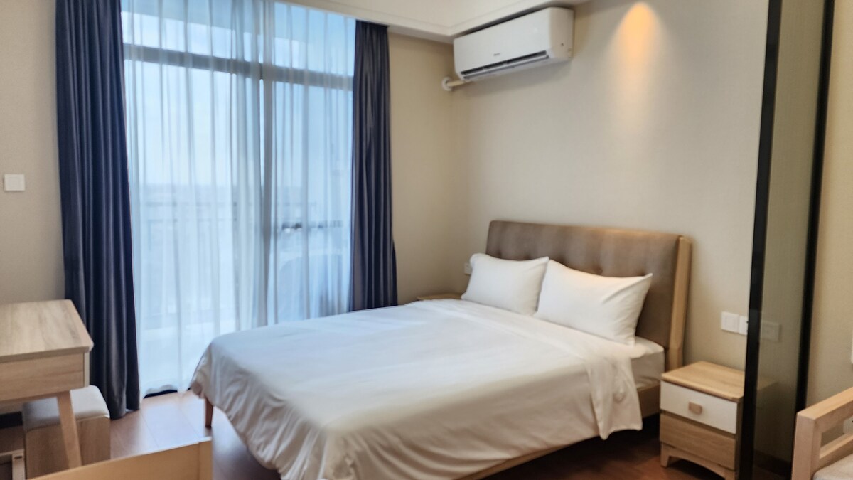 A well-lit bedroom features a spacious bed with crisp white linens and a soft headboard. A desk and chair are positioned nearby, with light filtering in through sheer curtains covering a large window, which provides a view of the city.