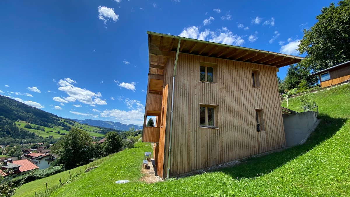 A wooden structure features a sloped roof and is surrounded by lush green grass. Large windows provide ample natural light, while the mountains and valley can be seen in the background under a clear blue sky. The environment conveys a sense of tranquility.
