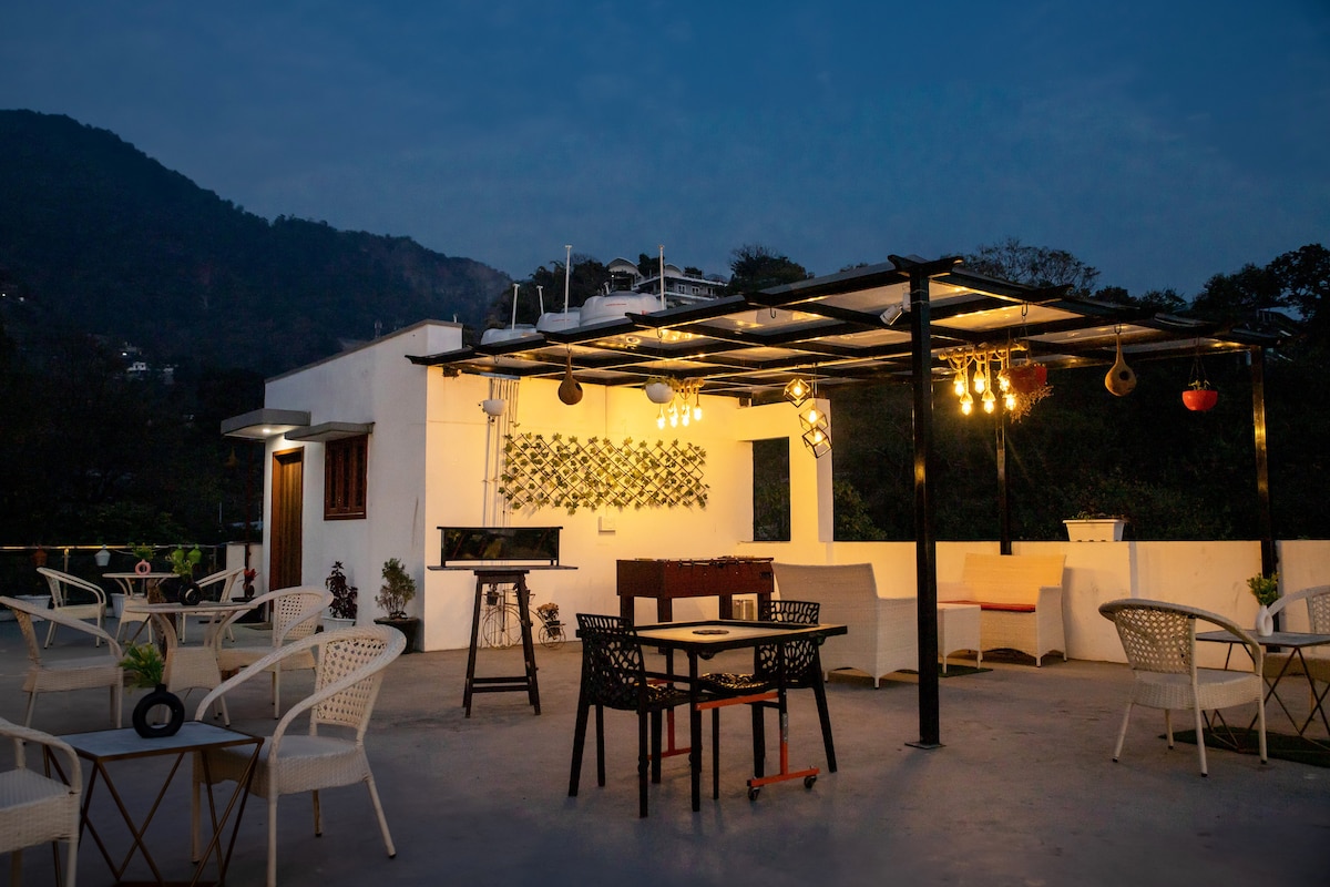 The rooftop seating area is showcased, featuring a variety of tables and chairs arranged under a canopy illuminated by soft lights. A barbecue grill is visible, with lush hills in the background, creating an inviting space for relaxation or social gatherings.