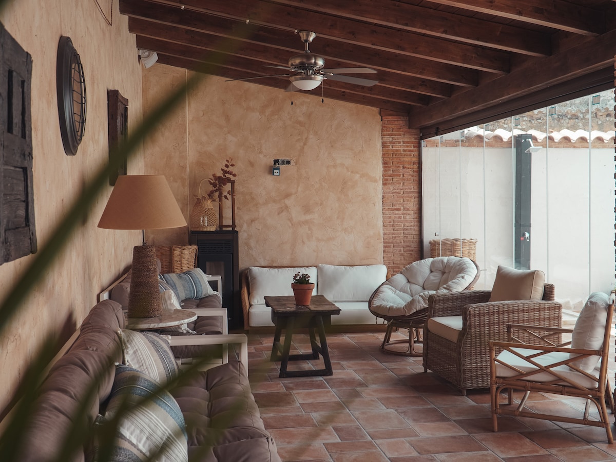 A spacious living area features a combination of seating options, including a white sofa and woven chairs. A small table sits in the center, while soft lighting from a lamp adds warmth. Large glass doors ensure abundant natural light and a view of the patio.