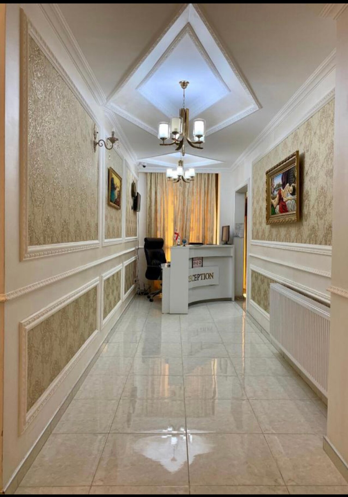 A well-lit hallway is visible, featuring elegant wall moldings and polished tiles. A reception desk is positioned at the end, backed by soft drapes filtering natural light. Artistic wall decor adds visual interest, and a chandelier overhead enhances the welcoming atmosphere.