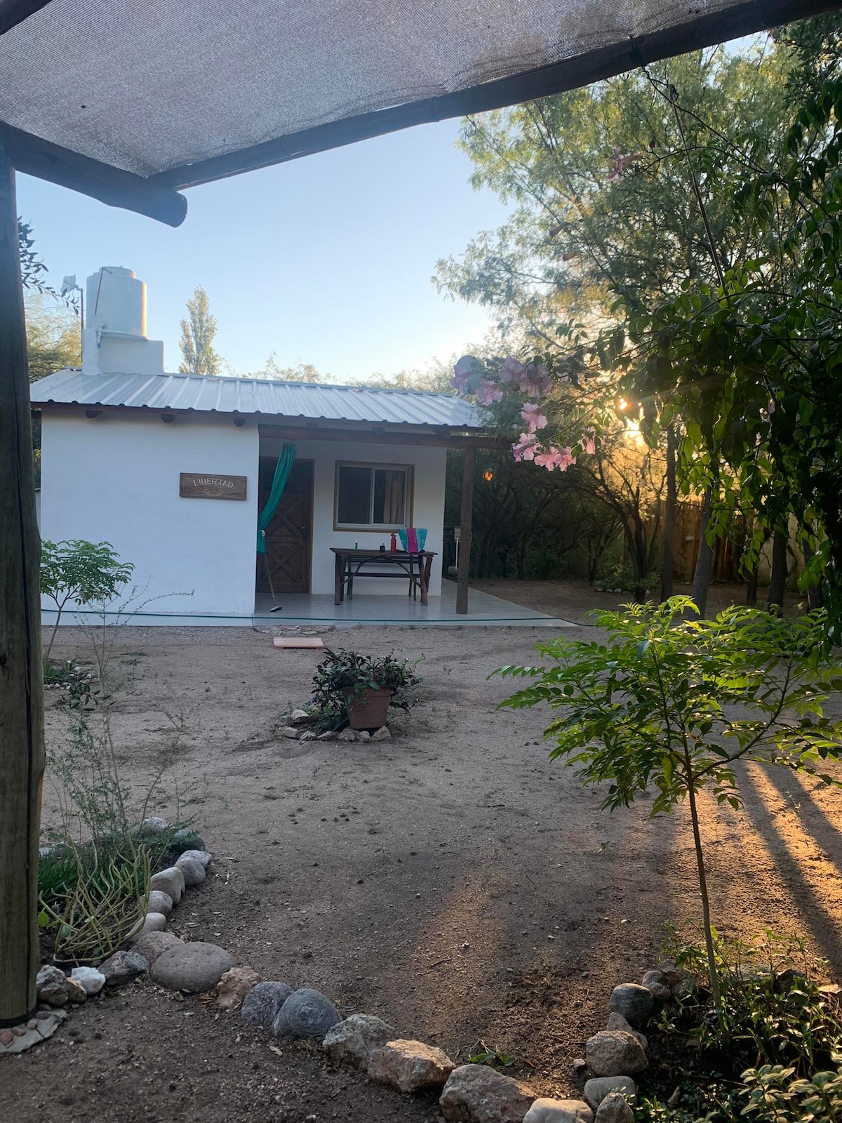 A charming accommodation is framed by lush greenery and flowered plants. The exterior features a simple white structure with a metal roof. A small outdoor dining area is visible, set up with a table and chairs, welcoming guests to enjoy the surrounding natural beauty.