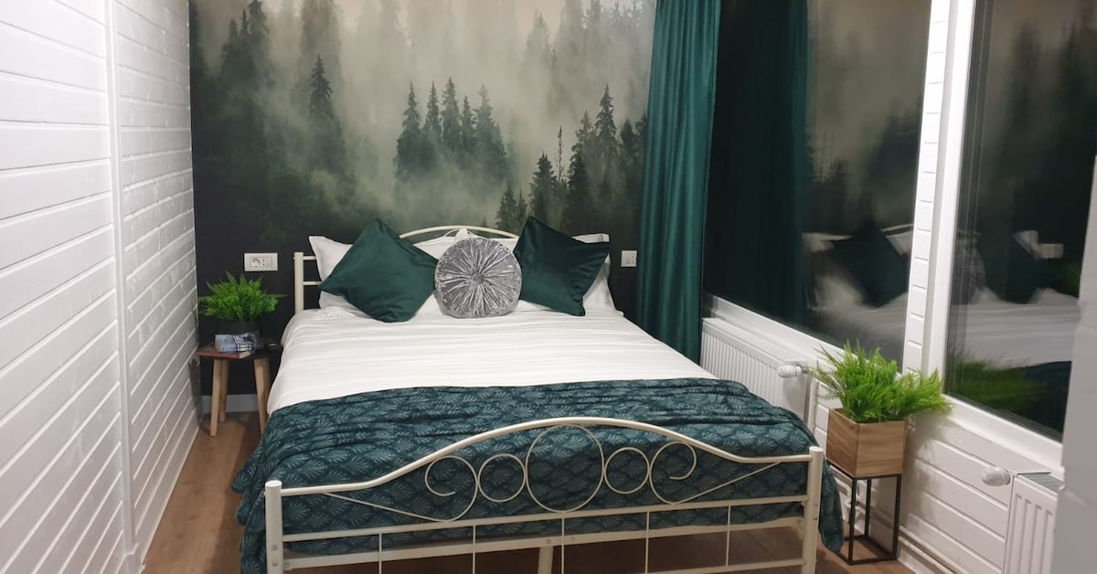 A cozy sleeping area features a bed with an elegant iron frame, adorned with a textured blue blanket and green accent pillows. Natural scenery wallpaper adds depth, while large windows with dark curtains frame the serene view, complemented by small potted plants on either side.
