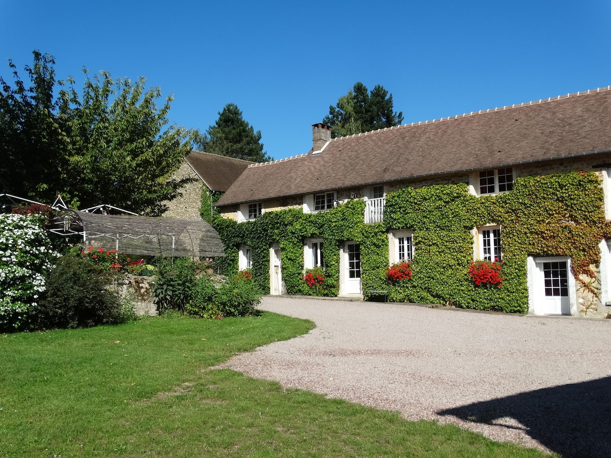 A charming farmhouse is presented, adorned with lush greenery and vibrant red flowers. The exterior features multiple windows, allowing for natural light. A gravel driveway leads to the entrance, set against a backdrop of neatly maintained lawns and surrounding trees.