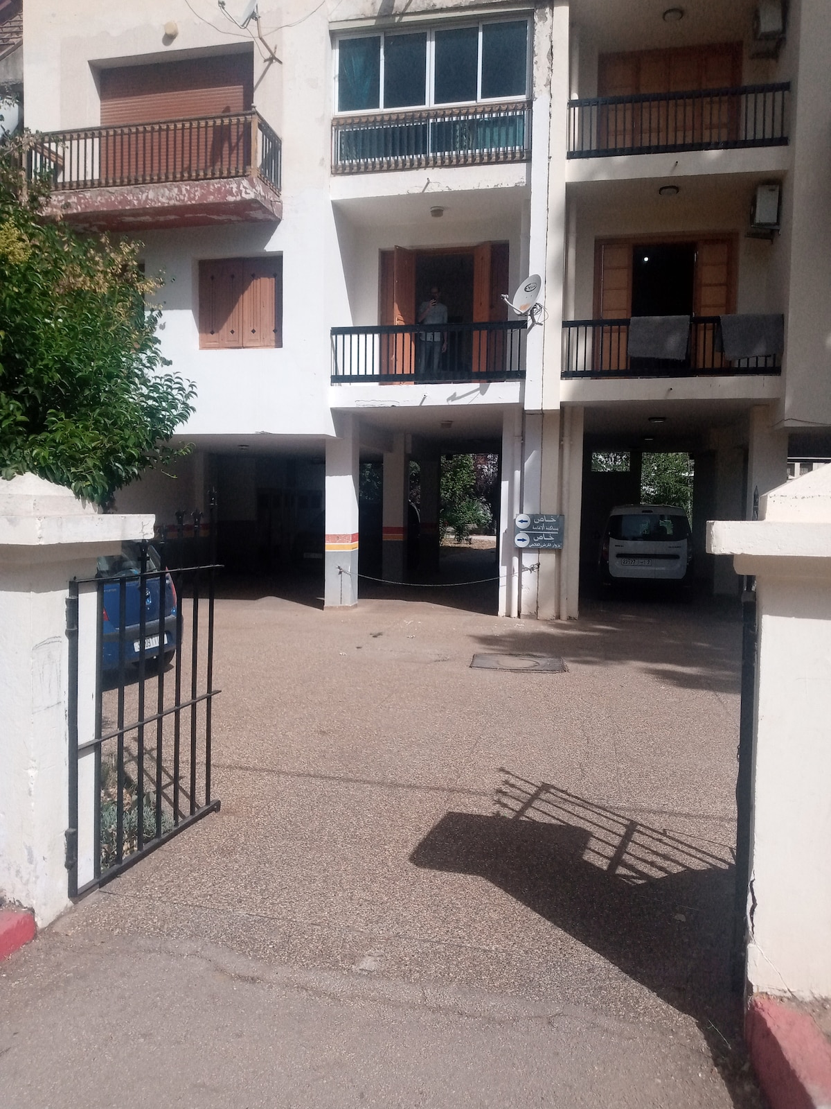 An open entrance leads to a spacious courtyard, flanked by two multi-story buildings. Balconies with black railings are visible on the upper floors. A blue vehicle is parked in the background, along with a white vehicle positioned nearby. Greenery adds a natural element to the surroundings.