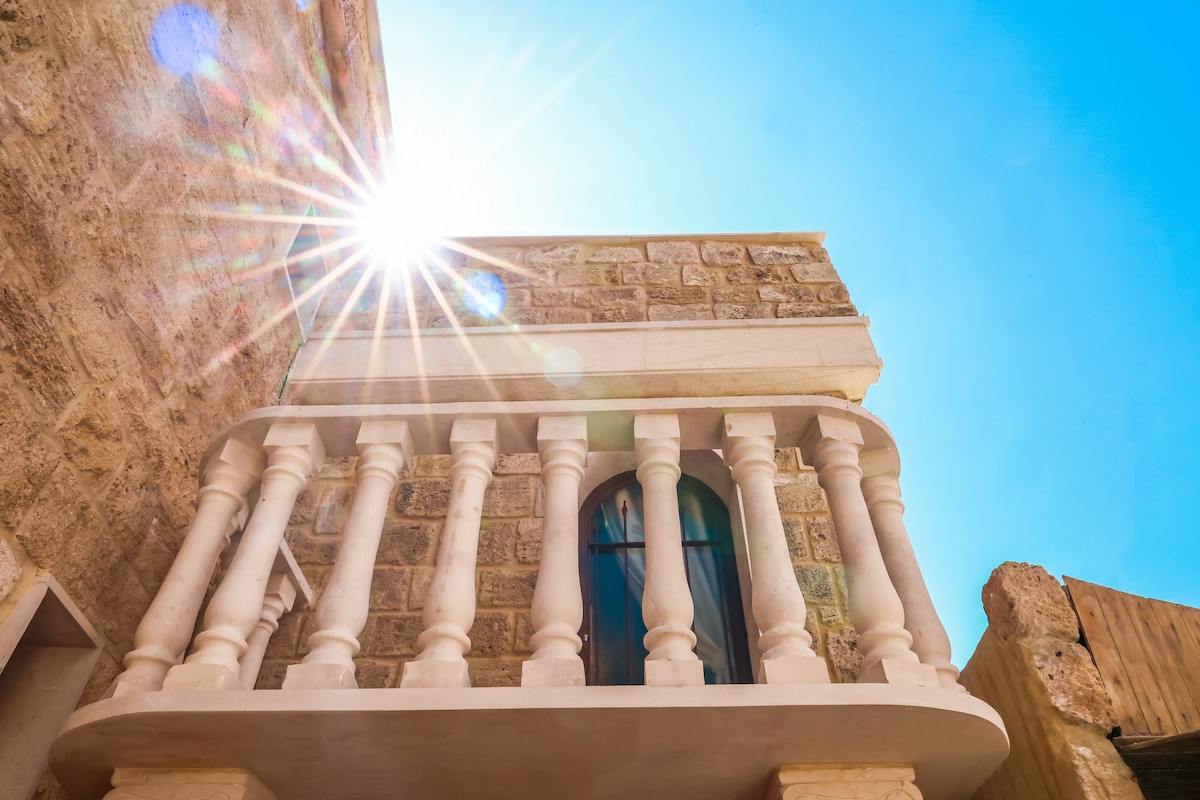 A small balcony is visible, featuring decorative columns and an arched window. The sun shines brightly above, illuminating the stone façade of the guesthouse, which showcases its historic charm. The clear blue sky serves as a backdrop, enhancing the inviting exterior.