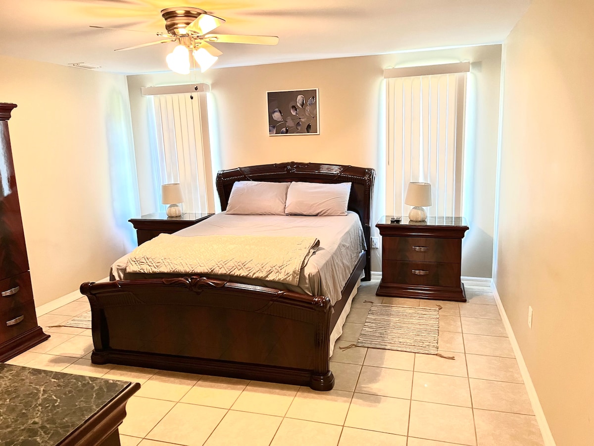 A spacious bedroom features a large bed with a light-colored quilt. Nightstands on either side hold table lamps. Natural light enters through two windows equipped with vertical blinds, while a soft rug is placed beside the bed on tiled flooring.